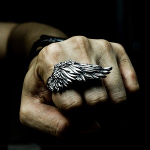 Peut inclure: Une bague en argent avec un motif d'aile détaillé. La bague est portée sur le majeur d'une main.