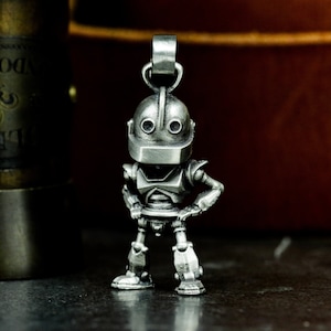 May include: A silver metal pendant shaped like a robot with a round head and two large eyes. The robot is standing with its hands on its hips.