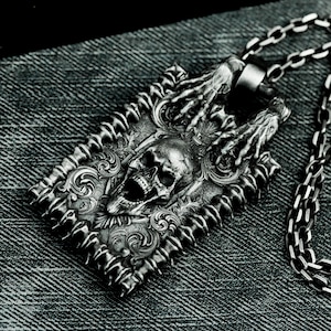 May include: A silver pendant necklace featuring a detailed skull design with skeletal hands reaching up. The pendant is rectangular and has a textured, ornate border. The chain is silver and has a box link design.
