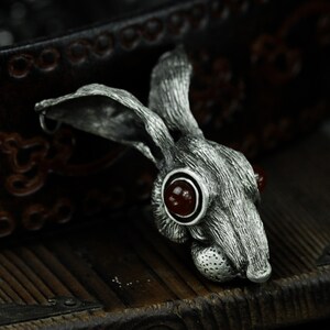May include: A silver rabbit pendant with red gemstone eyes. The pendant is detailed with fur texture and has a rustic, antique look.