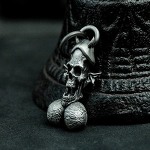 May include: A silver skull pendant with horns and two round balls attached to the bottom. The pendant is on a black surface.