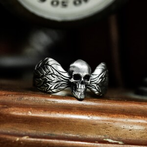 May include: A silver-colored ring featuring a skull with detailed wings. The skull has eye sockets, a nose cavity, and a jaw. The wings have a detailed feather design. The ring is set on a wooden surface.