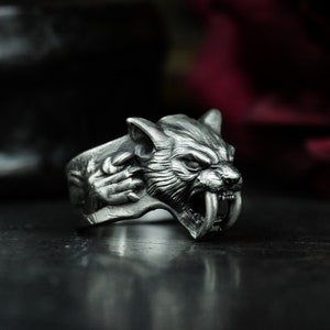 May include: A silver ring with a detailed design of a snarling saber-toothed tiger head. The ring is made of metal and has a realistic look.