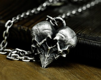 Puede incluir: Un collar con colgante plateado que presenta un diseño detallado de dos calaveras fusionadas. Las calaveras están intrincadamente esculpidas, con dientes y cuencas de los ojos visibles. La cadena del collar es de eslabones y el colgante descansa sobre madera.