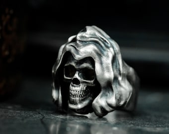 Op de afbeelding: Een zilveren ring met een gedetailleerd schedelontwerp dat een kapmantel draagt. De schedel heeft een gedetailleerd gezicht met een prominente neus en tanden.