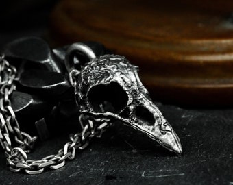 May include: A silver chain necklace with a detailed bird skull pendant. The pendant is textured and has a hollow eye socket.