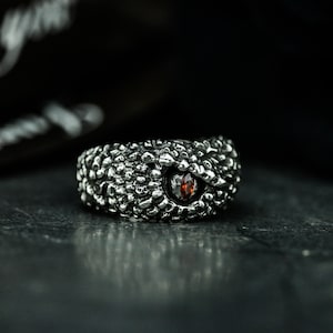 Op de afbeelding: Een zilveren ring met een getextureerd, schubbig ontwerp en een rode edelsteenoog.