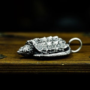 May include: A silver pendant in the shape of a snapping turtle. The turtle is detailed with a textured shell and a realistic head.