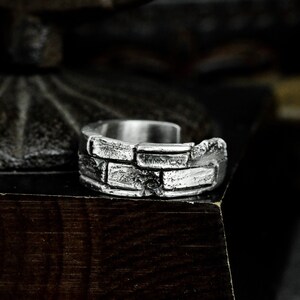 May include: A silver ring with a brick pattern design. The ring is made of metal and has a textured surface.