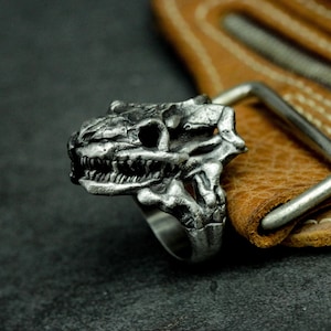 Op de afbeelding: Een zilveren ring met een gedetailleerd schedelontwerp. De schedel heeft een dreigende uitdrukking met scherpe tanden en een prominente kaaklijn. De ring is geplaatst tegen een bruine lederen achtergrond.