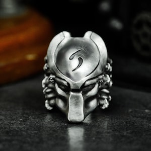 May include: A silver Predator-themed ring with a detailed sculpted head and a symbol on the forehead.