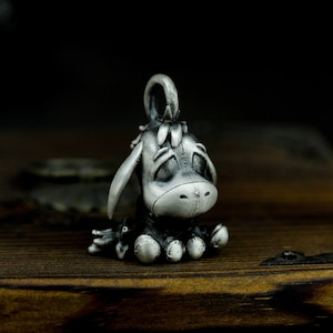 May include: A silver charm featuring a cartoon donkey with a sad expression. The charm is detailed with a textured surface and has a loop at the top for attaching to a necklace or bracelet.