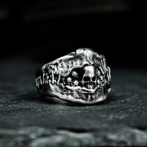 May include: A silver ring featuring a skull design. The skull is surrounded by a textured, organic-looking design. The ring is a statement piece with a gothic aesthetic.