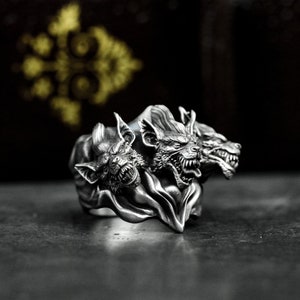 May include: A silver ring with three wolf heads, each with sharp teeth and a snarling expression. The heads are connected by a flowing, draped fabric.