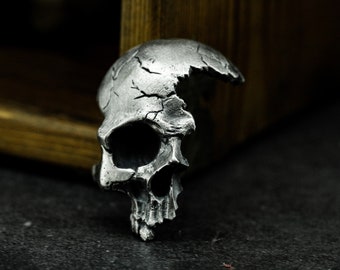 Könnte beinhalten: Ein silberner Totenkopfring mit einem rissigen und gebrochenen Design. Der Schädel ist detailliert mit einem markanten Unterkiefer und Augenhöhlen.