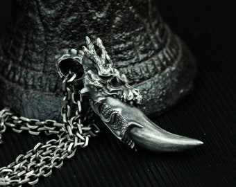 Puede incluir: Un collar de plata con un colgante de dragón con un diseño detallado. El colgante presenta una cabeza de dragón con dientes y cuernos afilados. El collar está hecho de una cadena de plata.