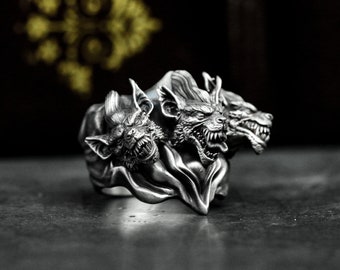 Könnte beinhalten: Ein silberner Ring mit drei Wolfsköpfen, jeder mit scharfen Zähnen und einem knurrenden Ausdruck. Die Köpfe sind durch einen fließenden, drapierten Stoff verbunden.