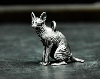 Puede incluir: Una figurita de plata detallada de un gato sin pelo que lleva un collar decorativo. El gato está sentado con la cola enroscada alrededor de sus patas. La figurita tiene un lazo en la parte superior, lo que sugiere que es un colgante.