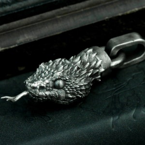 Op de afbeelding: Een zilveren slangenkopbedel met een getextureerd oppervlak en een gespleten tong. De bedel is bevestigd aan een zilveren ketting.