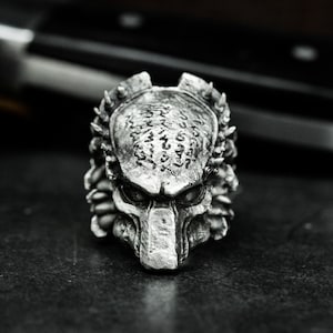 May include: A silver Predator head ring with intricate details and a textured surface. The ring features a detailed face with sharp teeth and a textured skull.