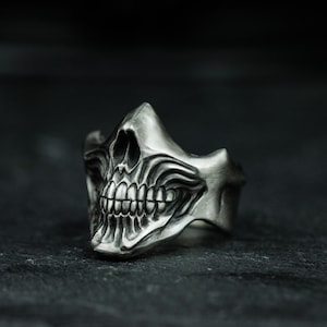 Könnte beinhalten: Ein silberner Totenkopfring mit einem detaillierten Design. Der Ring zeigt einen Totenkopf mit weit offenem Mund und scharfen Zähnen.
