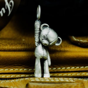 May include: A silver charm in the shape of a teddy bear with stitched details. The bear is standing on its hind legs with its arms raised above its head.