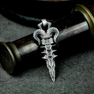 Może przedstawiać: Srebrny wisiorek w kształcie sztyletu z bogatym wzorem. Sztylet ma ostry czubek i ozdobny uchwyt z dwoma rogami barana. Wisiorek znajduje się na ciemnym tle.