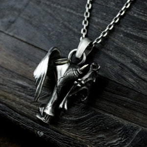 Puede incluir: Un collar con un colgante de plata que presenta una calavera detallada con un cuerno y un ala. La calavera sostiene una copa con un borde dorado.