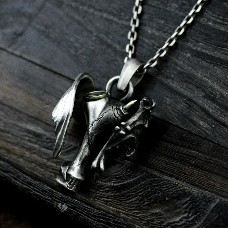 Puede incluir: Un collar con un colgante de plata que presenta una calavera detallada con un cuerno y un ala. La calavera sostiene una copa con un borde dorado.