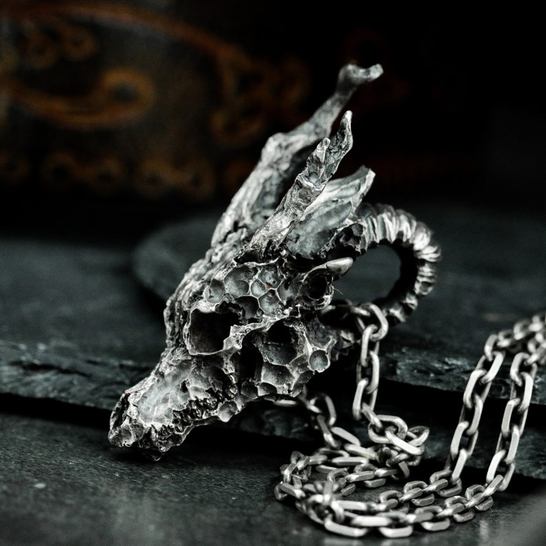 May include: A silver dragon skull pendant necklace with a detailed textured design. The pendant is attached to a silver chain.