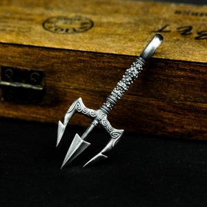 May include: A silver trident pendant with intricate details and a textured handle. The pendant is on a wooden surface.
