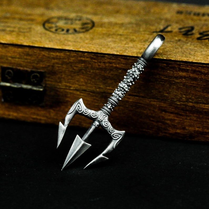 May include: A silver trident pendant with intricate details and a textured handle. The pendant is on a wooden surface.