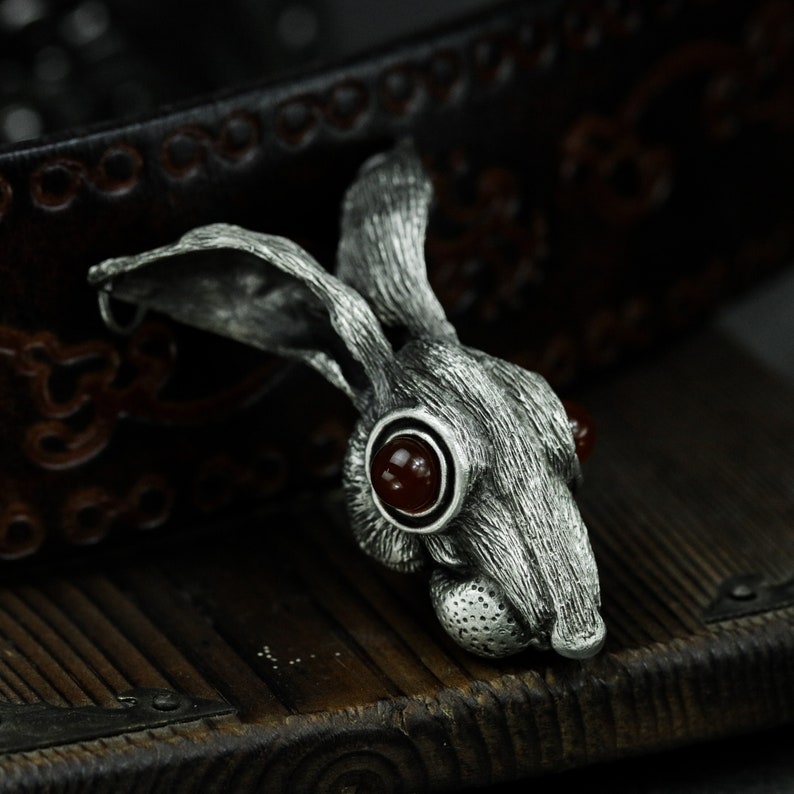 May include: A silver rabbit pendant with red gemstone eyes. The pendant is detailed with fur texture and has a rustic, antique look.