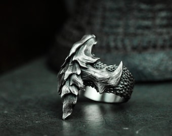 Puede incluir: Un anillo de plata con un diseño detallado de garra de dragón. La garra es texturizada y tiene puntas afiladas. El anillo está hecho de metal y tiene un acabado pulido.