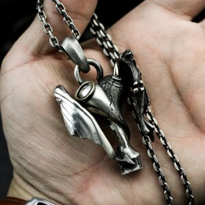 Puede incluir: Un collar con un colgante de plata que presenta un dise&ntilde;o tridimensional detallado de una criatura alada con un cuerno. El colgante est&aacute; unido a una cadena de plata.
