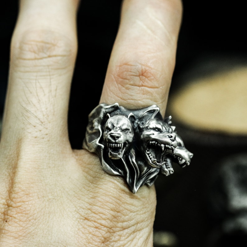 Puede incluir: Un anillo de metal plateado con un dise&ntilde;o detallado de tres cabezas caninas. Las cabezas est&aacute;n esculpidas con dientes expuestos y expresiones de gru&ntilde;ido, integradas en una banda texturizada con un acabado oscuro y oxidado.
