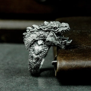 May include: A silver ring featuring a detailed dragon head design with an open mouth and sharp teeth. The dragon's scales and armour are intricately sculpted.