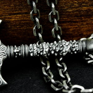 May include: A silver chain necklace with a detailed axe pendant. The axe has a textured handle and a sharp blade.