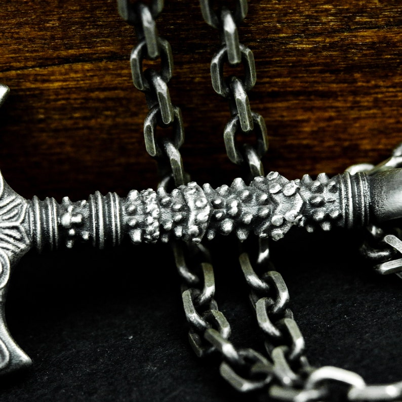 May include: A silver chain necklace with a detailed axe pendant. The axe has a textured handle and a sharp blade.