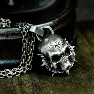 May include: A silver skull pendant necklace with a detailed design and a spiked halo. The necklace is made of a silver chain.