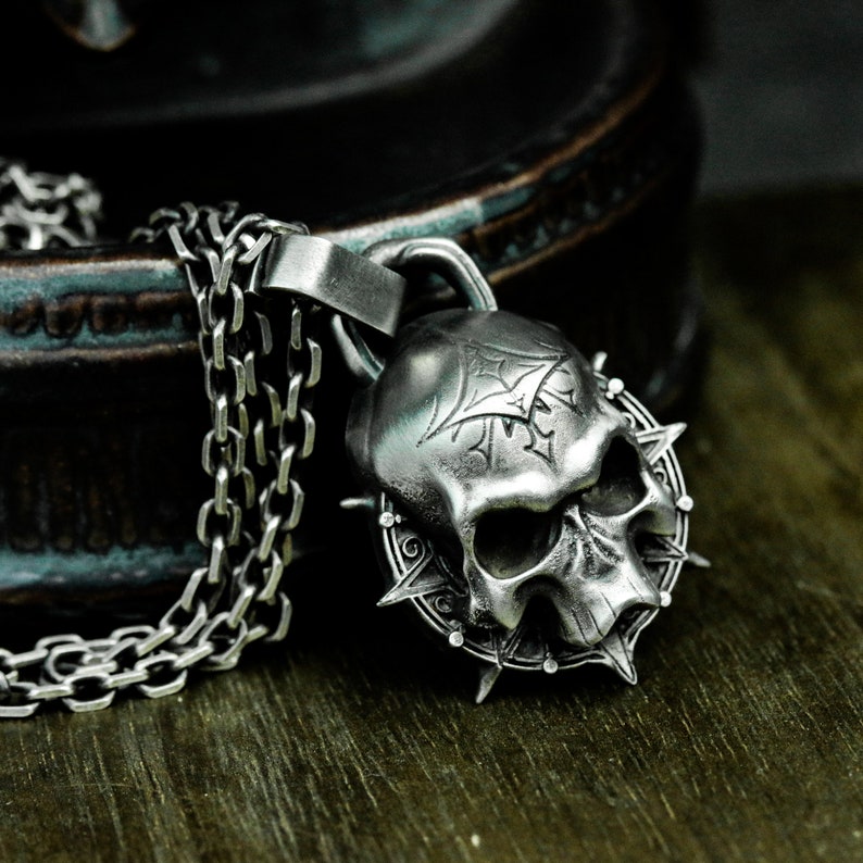 May include: A silver skull pendant necklace with a detailed design and a spiked halo. The necklace is made of a silver chain.