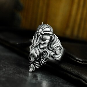 May include: A silver ring with a detailed, intricate design of a mythical creature's face. The ring is made of metal and has a textured surface.