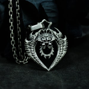 May include: A silver pendant necklace with a detailed design of two skulls with wings. The pendant is in the shape of a heart and has a chain attached.