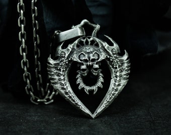 Puede incluir: Un collar con un colgante de plata con un diseño detallado de dos calaveras con alas. El colgante tiene forma de corazón y está unido a una cadena.