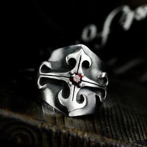 May include: A silver ring featuring an intricate cross design with a red gemstone at its centre.
