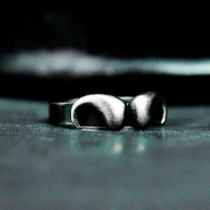 Può includere: Primo piano di un anello d'argento dal design unico. L'anello presenta due forme curve, simili a corna, collegate da una fascia. L'anello ha una finitura opaca ed è ambientato su uno sfondo scuro.