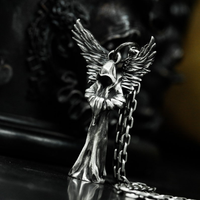 May include: A silver pendant featuring a hooded figure with wings. The figure is standing with its arms crossed and has a long, flowing robe. The pendant is attached to a silver chain.
