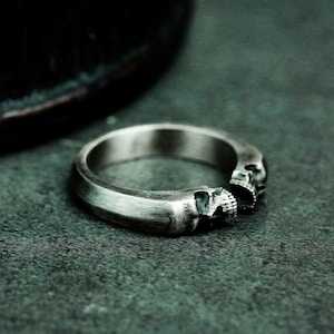 May include: A silver ring with two skull designs on the band. The ring is on a dark gray surface.