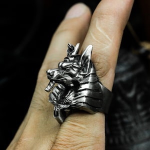 Może przedstawiać: Srebrny pierścionek z detalicznym wzorem głowy Anubisa. Pierścionek jest noszony na palcu.