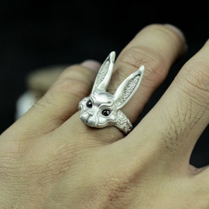 May include: A silver ring with a detailed rabbit head design. The rabbit's ears are long and pointed, and its eyes are black. The ring is worn on a finger.